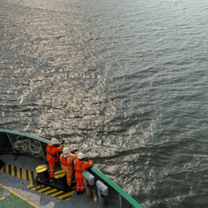 Three seafarers in orange safety suits observing sea surface from ship deck, symbolizing situational awareness and safety culture onboard vessels.
